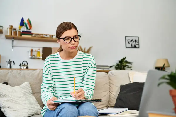 Una joven mujer hermosa se sienta en un sofá, tomando notas con atención mientras mira su computadora portátil. - foto de stock