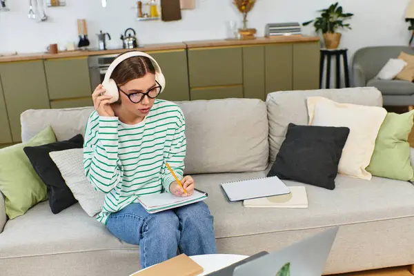 In a cozy living space, a young woman wears headphones, focused on her studies with notepad ready. — Stock Photo