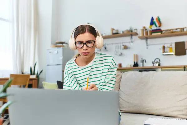 Focalizzato giovane donna con le cuffie prende appunti mentre studia sul suo computer portatile in uno spazio luminoso — Foto stock