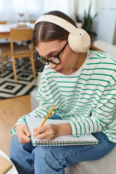 A young woman with headphones sits on a couch, absorbed in writing in her notebook. — Stock Photo