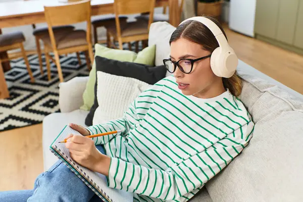Une jeune femme avec écouteurs prend des notes dans son confortable salon meublé en bois. — Photo de stock