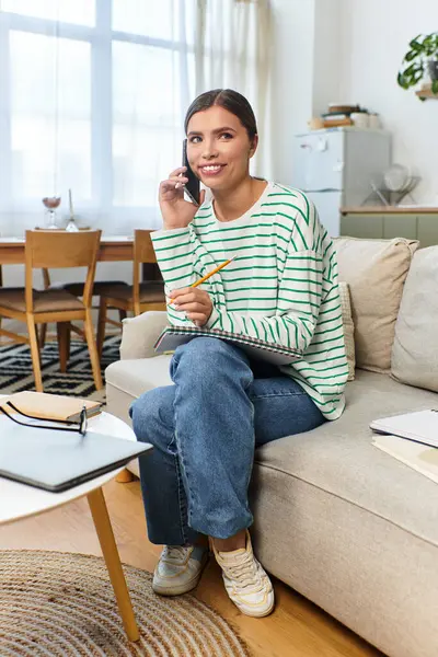 A young woman smiles as she talks on the phone, jotting notes in a modern and cozy living room. — Stock Photo