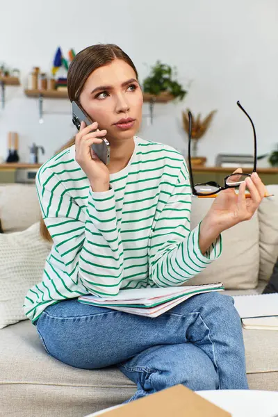 A young woman sits on a sofa, thoughtfully talking on the phone amid study materials. — Stock Photo