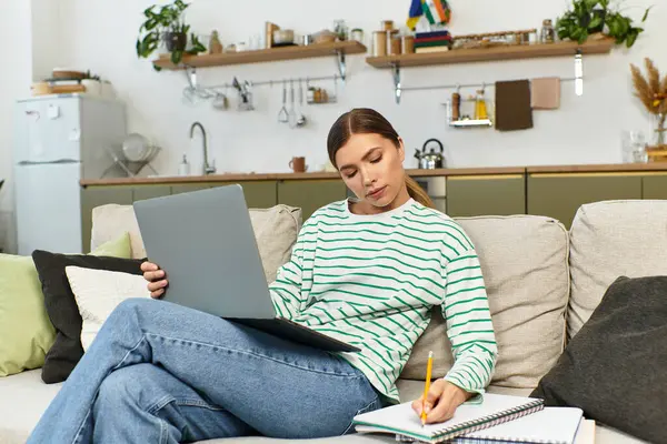 A young woman relaxes on a couch, focused on her laptop and jotting notes. — Stock Photo