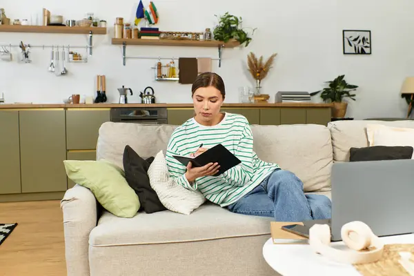 A young woman sits comfortably on a sofa, writing her thoughts in a notebook. — Stock Photo