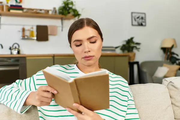 Femme en pull rayé lit un livre tout en se relaxant sur un canapé doux dans une pièce lumineuse. — Photo de stock