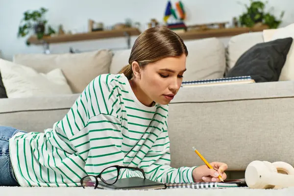 Una joven con una camisa a rayas escribe en su cuaderno en el suelo de la sala de estar. - foto de stock