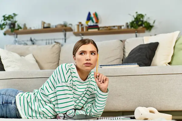 A beautiful young woman lies on her stomach, pondering while surrounded by books in a stylish room. — Stock Photo