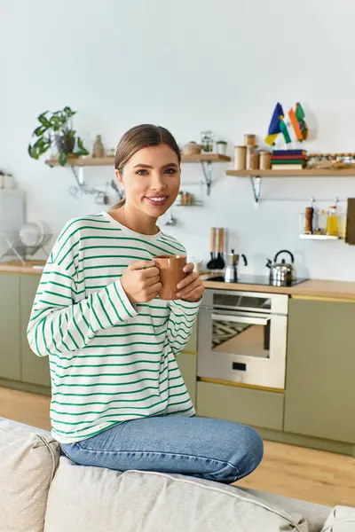 A young beautiful woman sits on a couch, sipping coffee while relaxing in her stylish kitchen. — Stock Photo