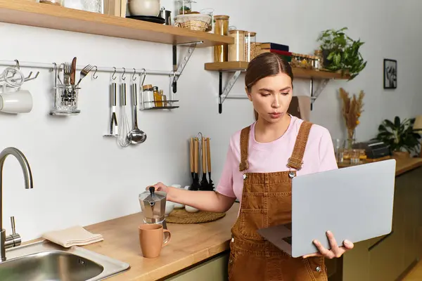 A young beautiful woman is preparing coffee while multitasking on her laptop in a stylish kitchen. — Stock Photo