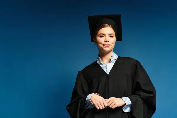 Joyful young woman proudly stands wearing a graduation cap and gown against a blue backdrop. — Stock Photo