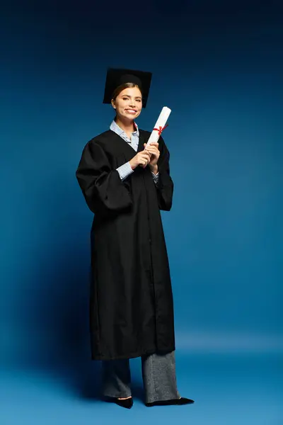 In a bright blue background, a joyful young woman in graduation robes holds her diploma. — Stock Photo