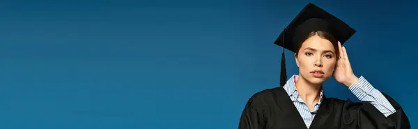 Молодая женщина в выпускном платье и кепке элегантно позирует на синем фоне, демонстрируя гордость. — стоковое фото