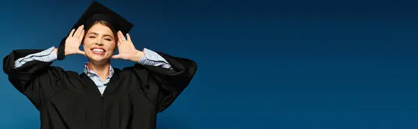 Happy graduate in cap and gown smiles, celebrating her success against a blue background. — Stock Photo
