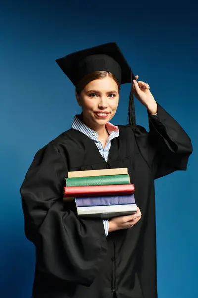 Young beautiful woman in graduation cap proudly holding a stack of colorful books, smiling joyfully — Stock Photo