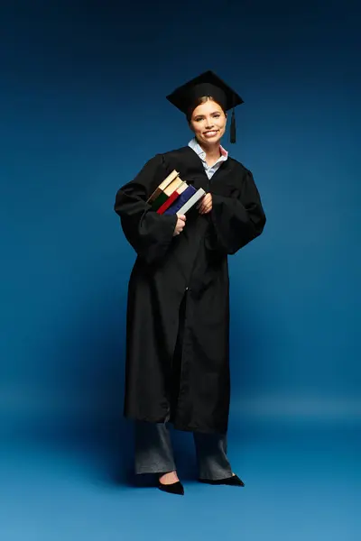 Giovane donna in abito da laurea e cappello tiene libri mentre sorride su uno sfondo blu. — Foto stock