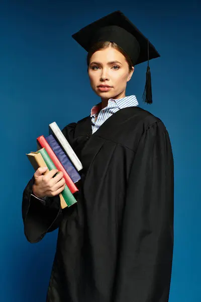 Una joven orgullosa en un vestido de graduación sostiene libros, radiante sobre un telón de fondo azul. — Stock Photo
