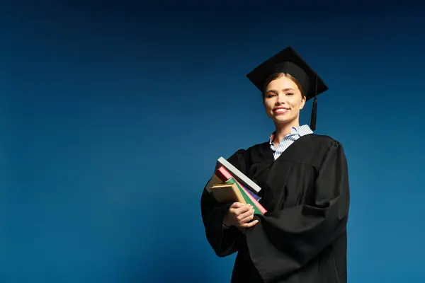 A joyful young woman celebrates her graduation in a stylish cap and gown while holding books. — Stock Photo