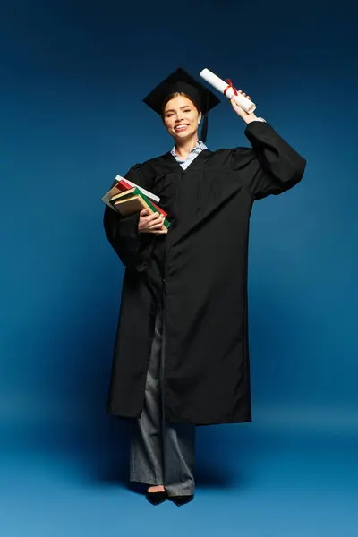 Bright smile on a young beautiful woman in graduation attire holding diploma and books excitedly. — Stock Photo