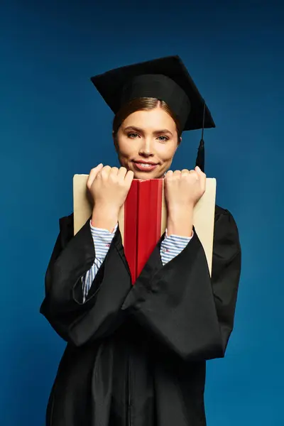 Young beautiful woman proudly holds a red book while dressed in her graduation cap and gown. — Stock Photo