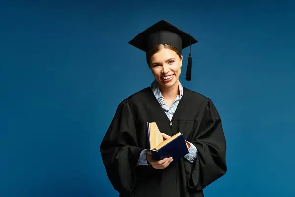 A young woman in a cap and gown smiles broadly while holding books against a blue background. — Stock Photo