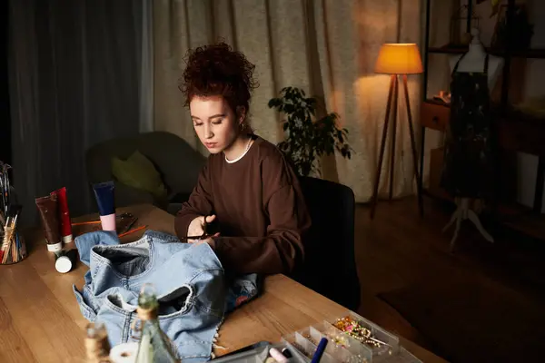 A skilled craftswoman designs a unique denim jacket in a cozy, artful space during the evening. — Stock Photo