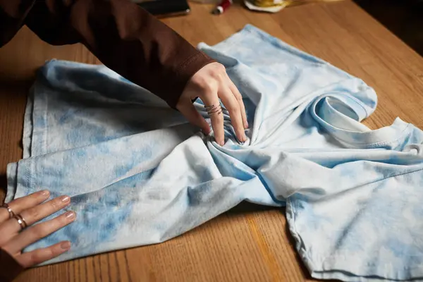 Young craftswoman skillfully designs a tie dye pattern on a light blue shirt in her workspace. — Stock Photo