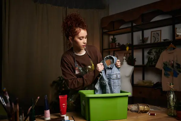 Junge Kunsthandwerkerin taucht Jeansjacke in leuchtend grünen Container, umgeben von Kunstgegenständen. — Stockfoto