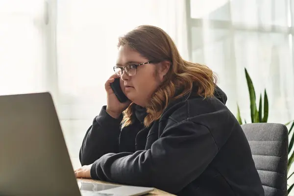 In a cozy home, a young woman with Down syndrome chats on the phone while using her laptop. — Stock Photo