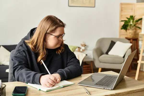 A young woman with Down syndrome writes in her notebook, focused on her laptop at home. — Stock Photo