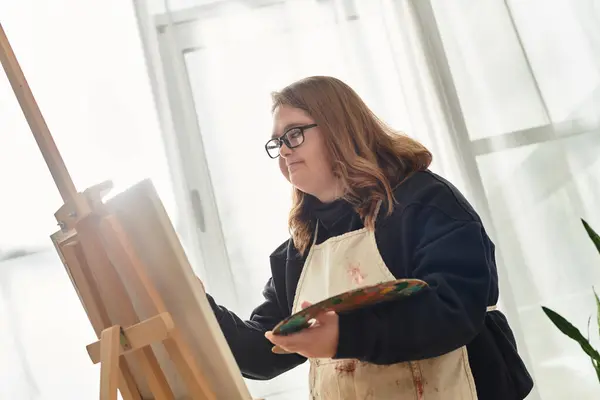 A young woman with Down syndrome joyfully paints on canvas at home, embracing her creativity. — Stock Photo