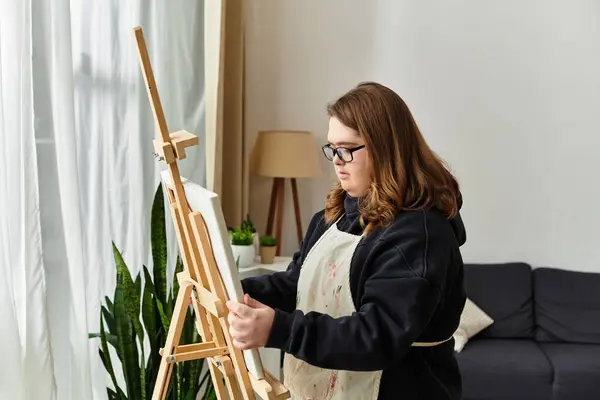A confident young woman with Down syndrome paints on an easel while dressed comfortably at home. — Stock Photo