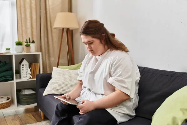 A young woman with Down syndrome relaxes at home, focused on her tablet in casual clothes. — Stock Photo