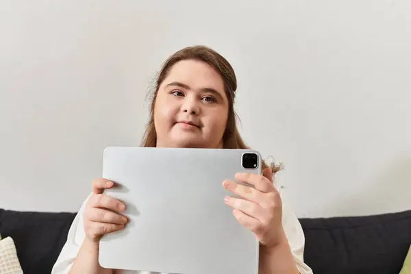 A young woman with Down syndrome sits relaxed at home, holding a tablet while exuding confidence. — Stock Photo