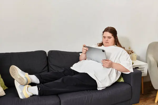 A young woman with Down syndrome lounges comfortably on a couch at home, focused on her tablet. — Stock Photo