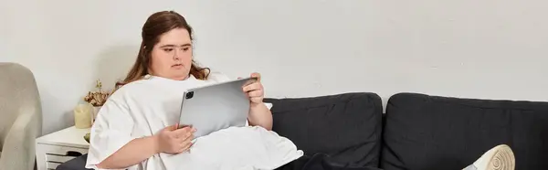Young woman with Down syndrome relaxes comfortably at home, engaged with a tablet while seated. — Stock Photo