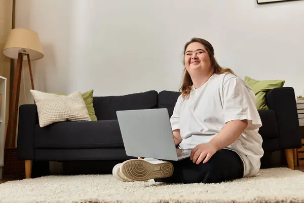 A young woman with Down syndrome sits comfortably on the floor, smiling as she uses her laptop. — Stock Photo