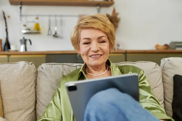 A joyful senior woman uses her tablet while relaxing in a chic living room. — Stock Photo