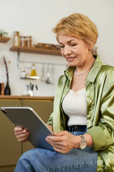 Beautiful mature woman explores digital content on her tablet, smiling with joy in a cozy kitchen. — Stock Photo
