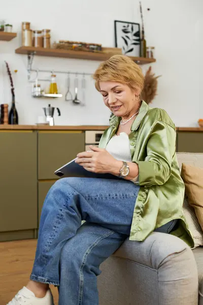 A joyful mature woman relaxes at home, reading on her tablet. — Stock Photo