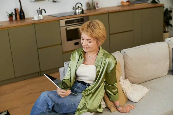 Beautiful senior woman in a green blouse relaxes on a couch while reading a book in her home. — Stock Photo