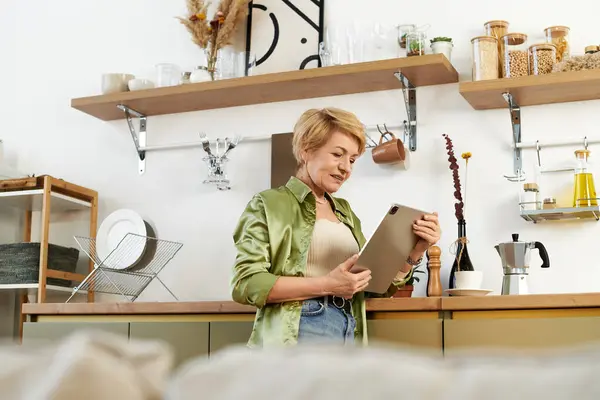 Eine schöne Seniorin steht mit ihrem Tablet in einer hellen Küche voller charmanter Dekoration. — Stockfoto