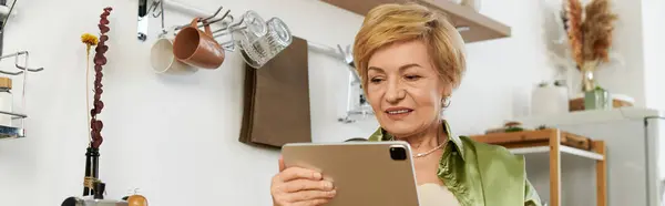 Красивая пожилая женщина улыбается, исследуя свой планшет в теплой и уютной кухне. — стоковое фото