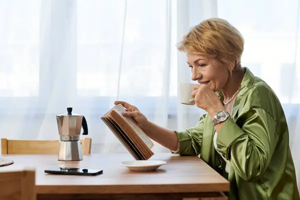 Beautiful senior woman sipping coffee while reading a book at a cozy kitchen table in daylight — Stock Photo