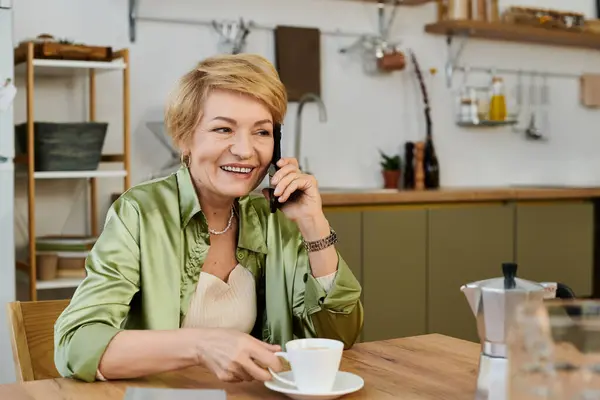 A beautiful senior woman talks on the phone, smiling while having coffee in her inviting kitchen. — Stock Photo