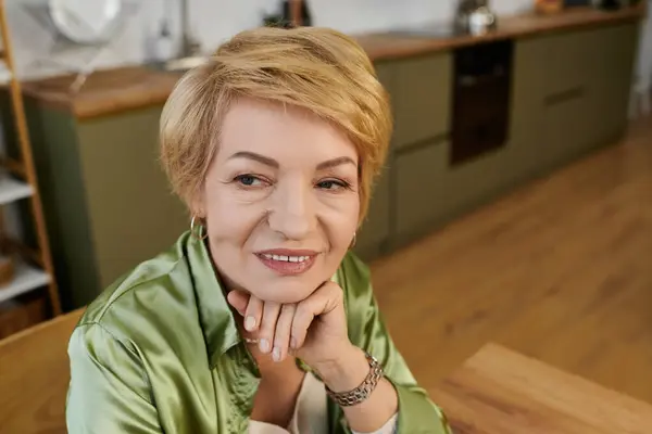 Senior woman with a thoughtful expression sits in a stylish kitchen, exuding grace and warmth. — Stock Photo