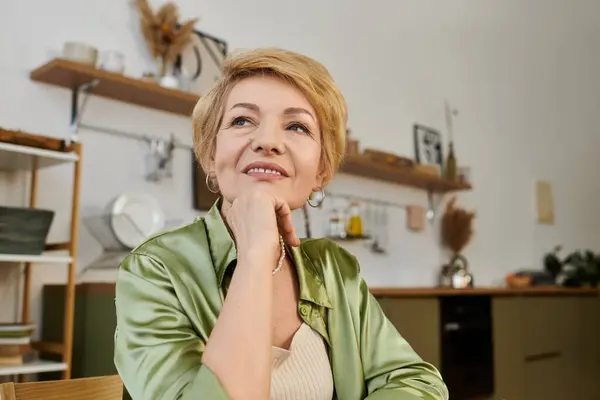 Красивая пожилая женщина задумчиво сидит на оживленной кухне, наслаждаясь моментом размышлений. — стоковое фото