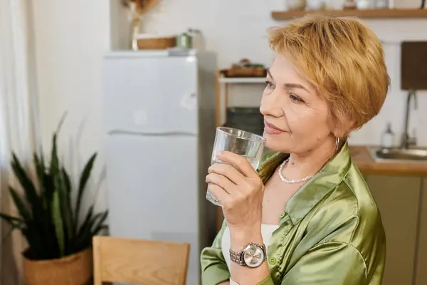 Mujer madura con el pelo corto y rubio sonríe mientras saborea una bebida en un espacio de cocina brillante. - foto de stock