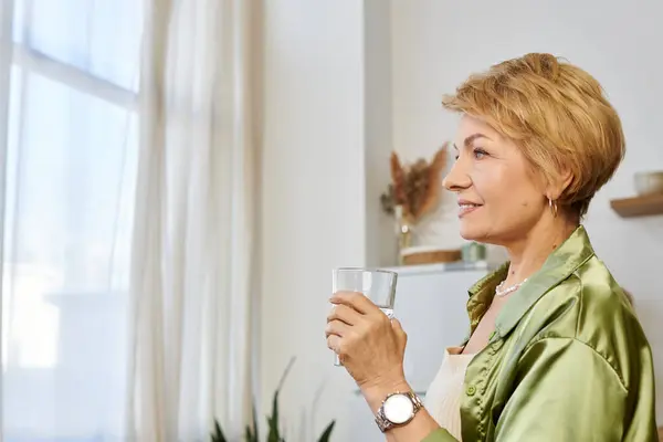 Una encantadora mujer mayor con el pelo corto sorbe agua, disfrutando de un momento tranquilo junto a la ventana. - foto de stock