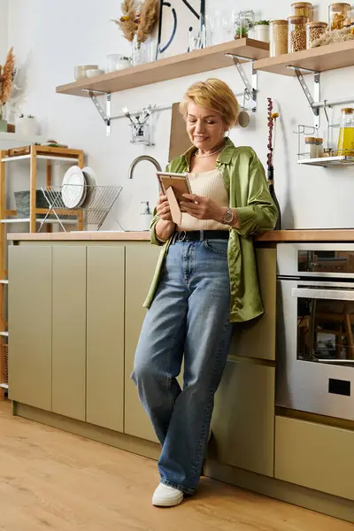 Senior woman stands in a stylish kitchen, reading a book, radiating happiness and contentment. — Stock Photo